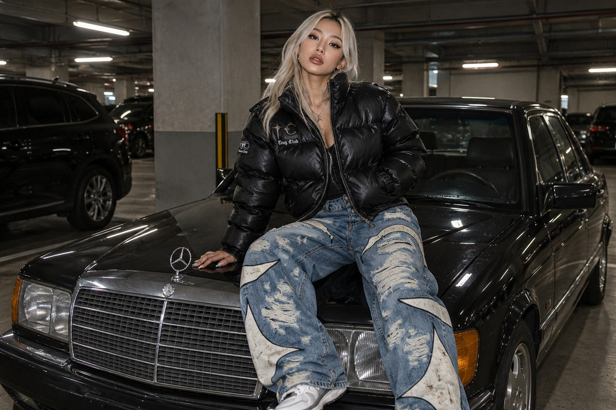 Woman in a black leather puffer jacket and washed jeans sitting on an old black Mercedes in an underground car park