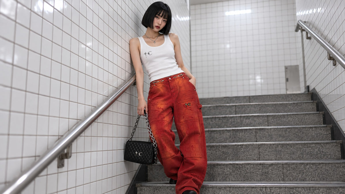 Woman in a white tank top and red washed pants holding a quilted chain bag in a white-tiled stairwell