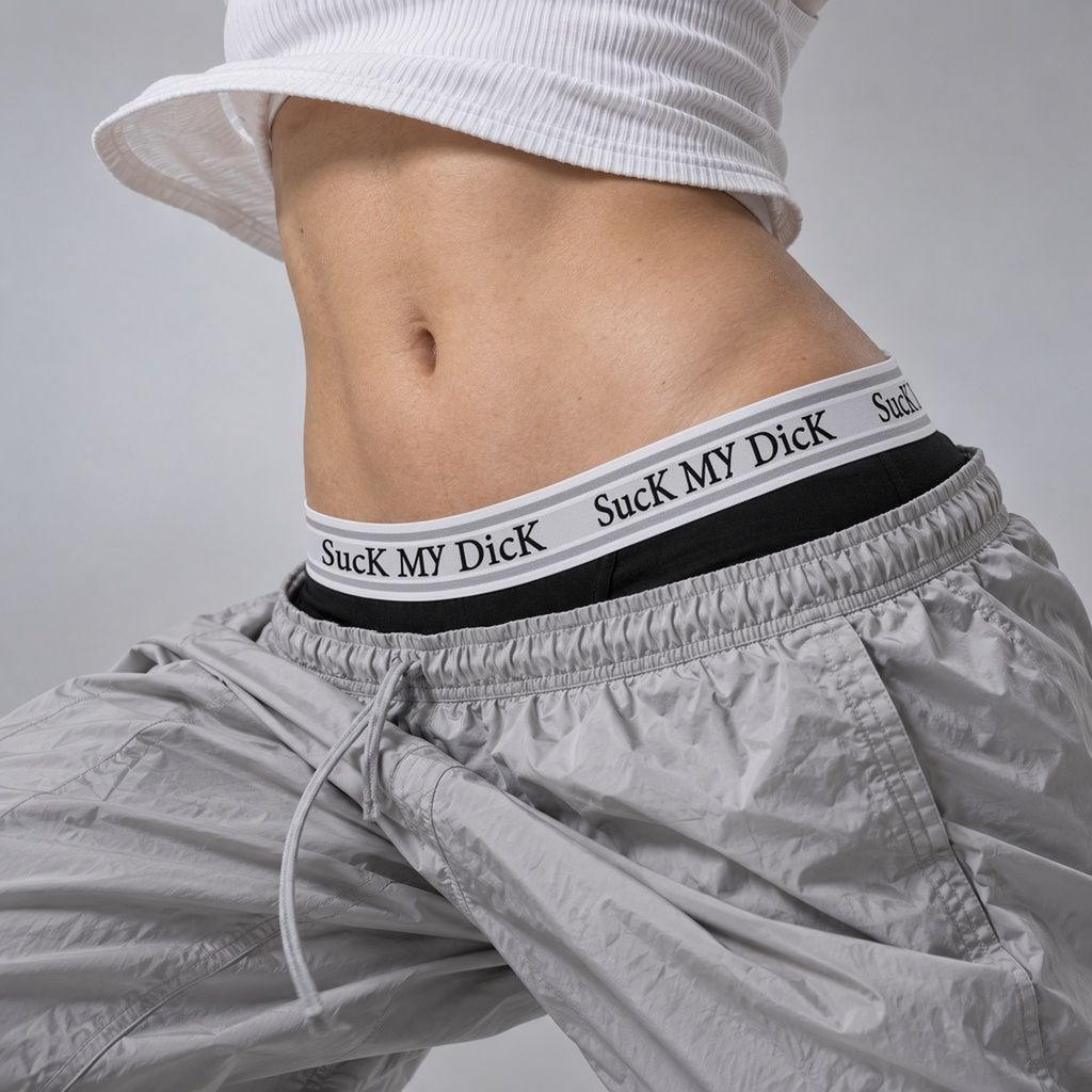 Woman wearing black Suck My Dick boxer briefs with a white and grey waistband, worn low under grey nylon track pants with a cropped white ribbed tank top